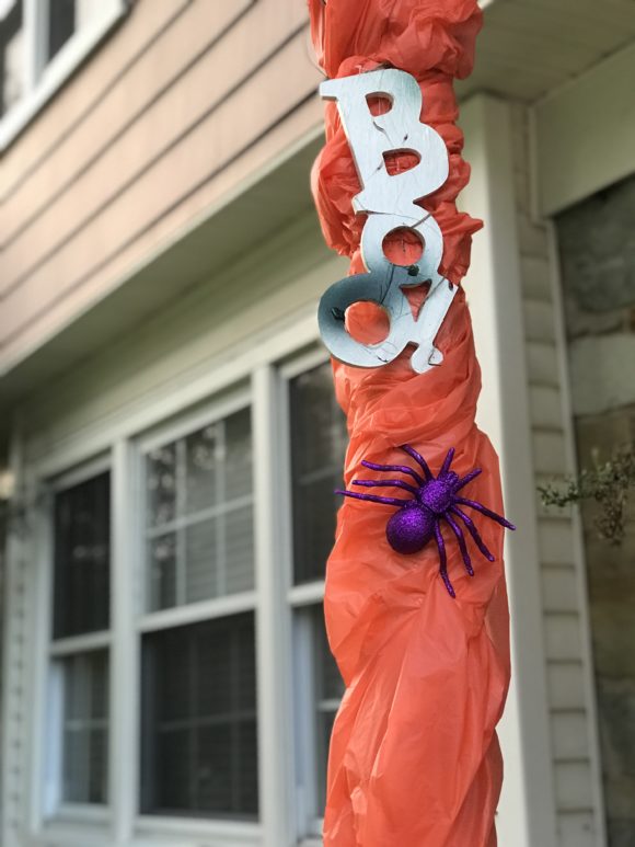 A front porch column is decorated for Halloween with a boo sign, spider and an orange background.
