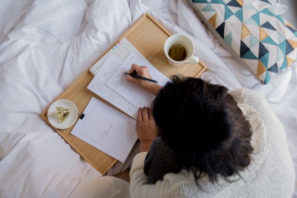 Person writing travel plans for a road trip in journal while sitting on a bed.