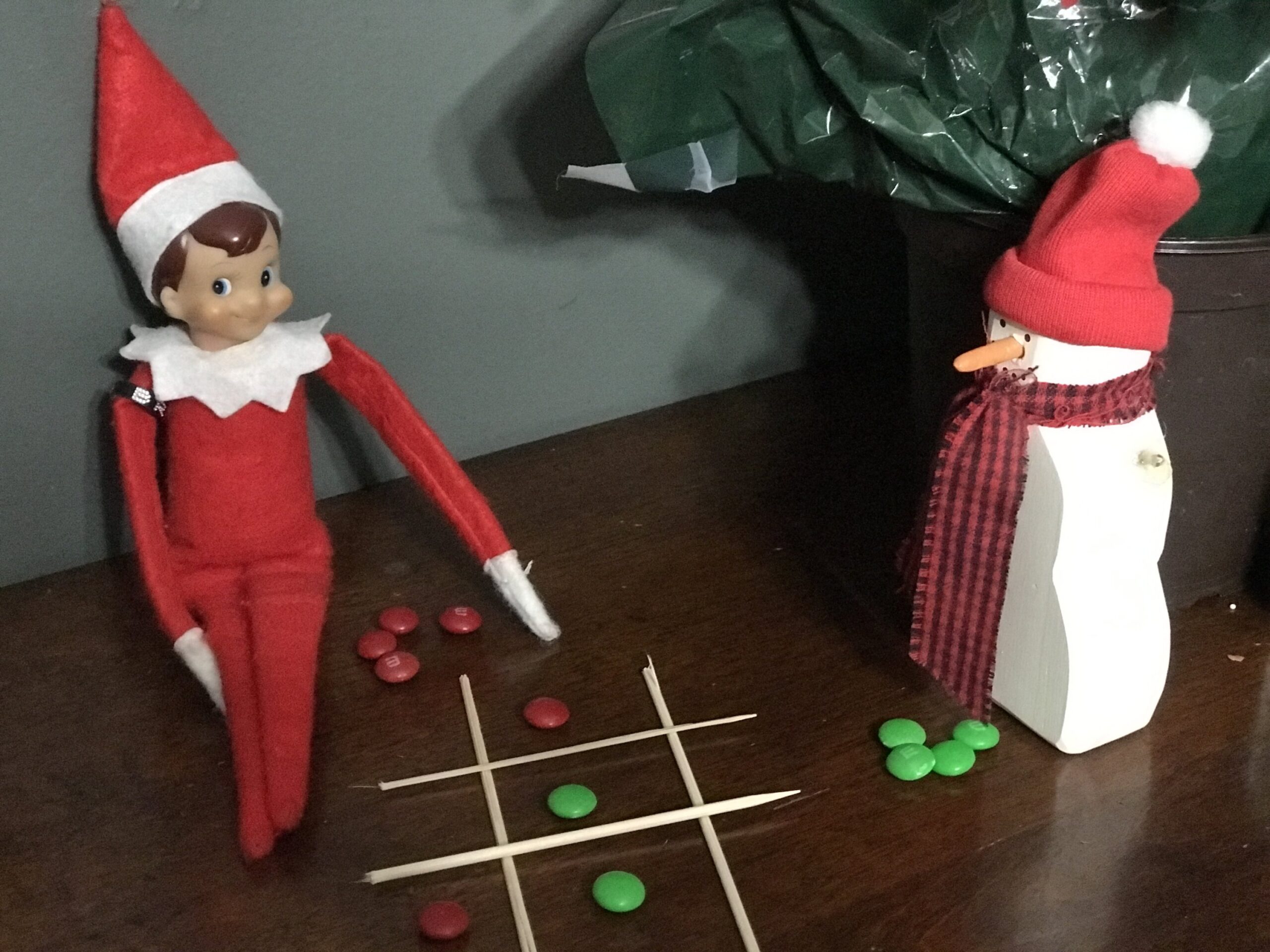 Elf on the Shelf playing tic tac toe with m&ms and toothpicks with a small wooden snowflake