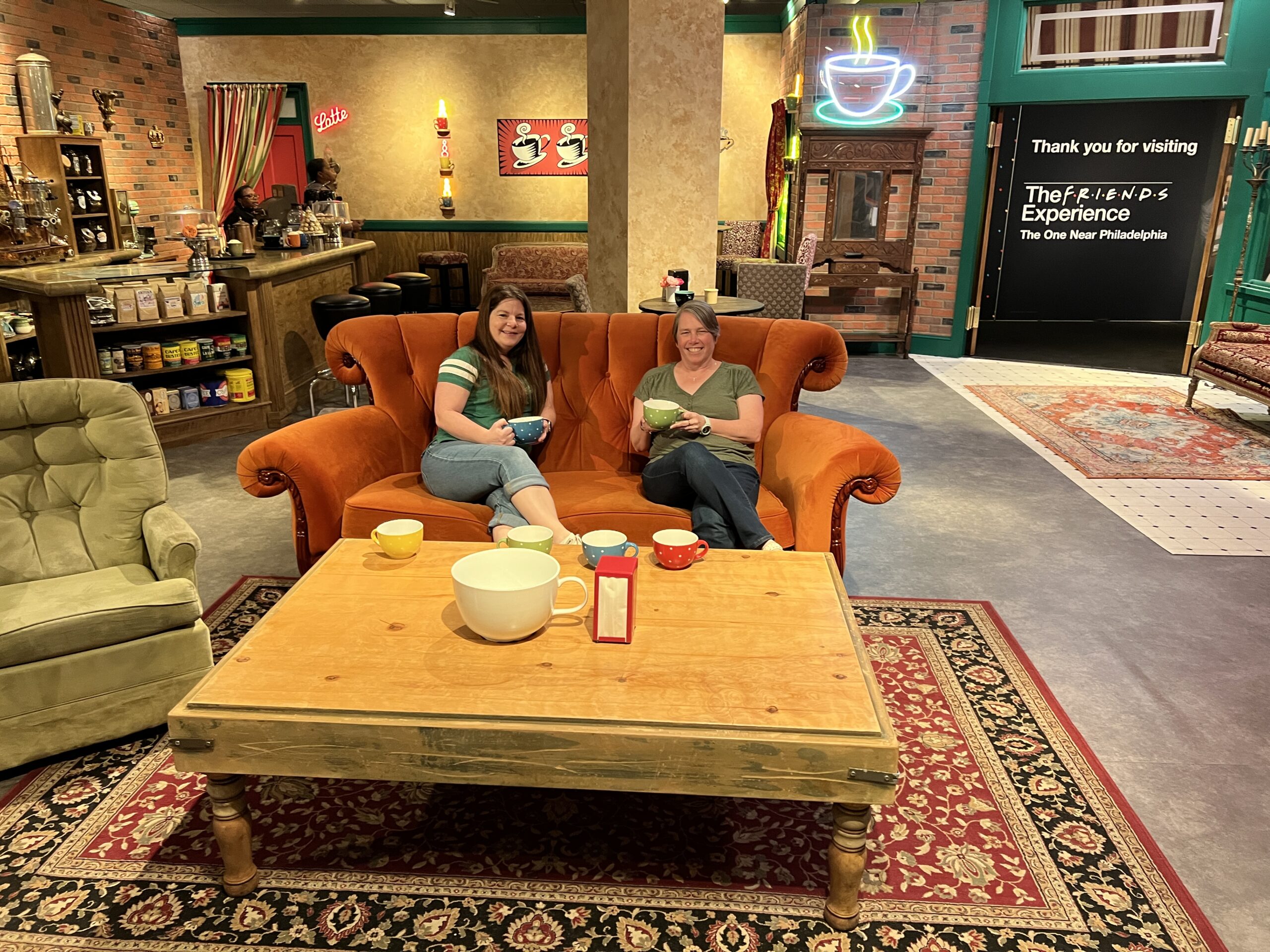 Kathy and Jenn at Central Perk on Orange Couch with Coffee mugs nonbranded image at The Friends Experience Near Philadelphia